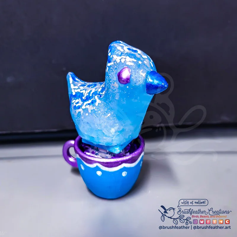 A hand cast resin clicking bird in a tiny teacup. This one is a translucent blue painted with ripples and waves along its back. The beak is a shiny darker blue and the eye is a solid shiny purple. The cup is painted in purple, white and blue with a curtained edge.