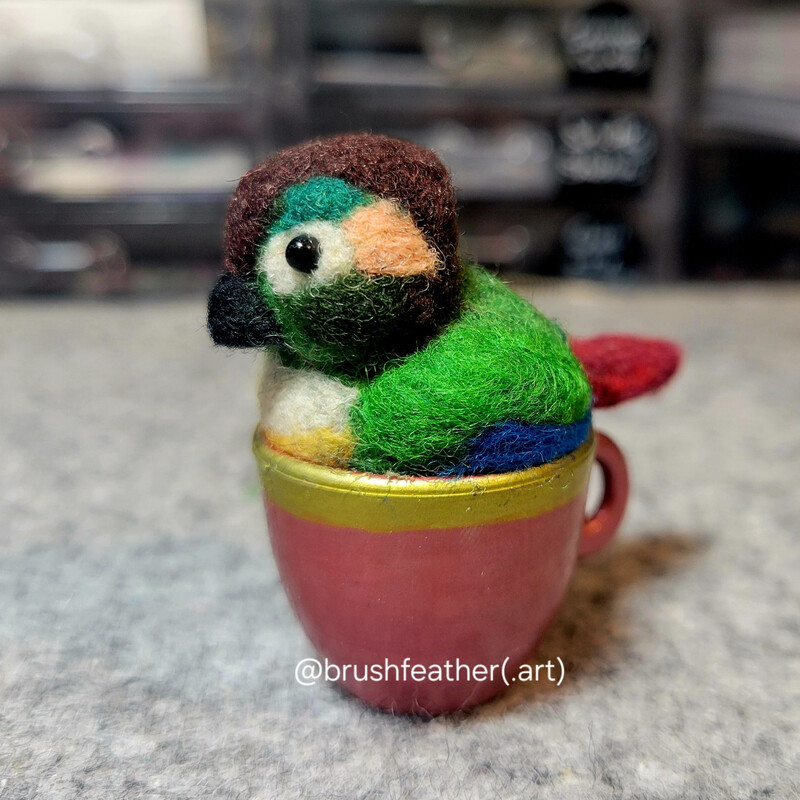 A needle felted green cheek conure sitting in a red and gold teacup. It's about 2
